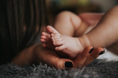 The baby's feet are holding a woman's hand, and manicured hands over paint on the grey blanket
